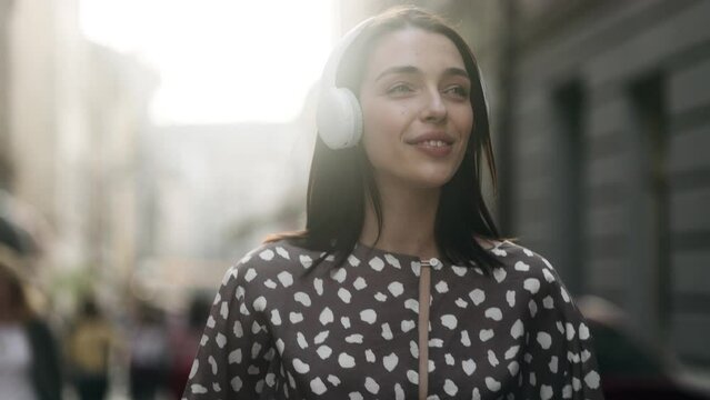 Attractive young woman listening to music in headphones and walking down the city street. Portrait of beautiful brunette.