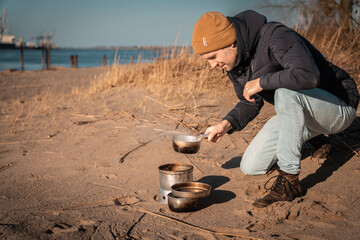Young white man an his knees with warm clothes on is cooking with outdoor cooker on Harriersand...