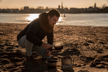 Young white man an his knees with warm clothes on is cooking with outdoor cooker on Harriersand...