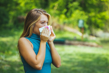 Young pretty woman blowing nose in front of blooming tree. Spring allergy concept. Attractive young adult woman coughing and sneezing outdoors. Sick people allergy or virus influenca concept.