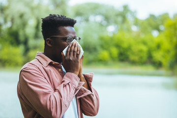 Man with allergy or an infection sneezing. Shot of a young man blowing his nose outdoors. His...