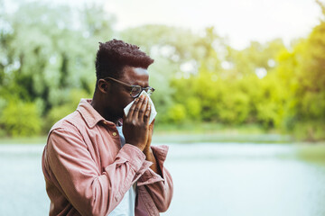 Man with allergy or an infection sneezing. Shot of a young man blowing his nose outdoors. His immune system needs a booster. Hipster man Sneezing. - obrazy, fototapety, plakaty