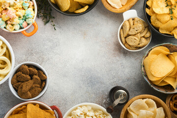 Snacks. Unhealthy food. All classic potato snacks with peanuts, popcorn and onion rings and salted pretzels in bowl plates on gray concrete background. Unhealthy food for figure, heart, skin, teeth