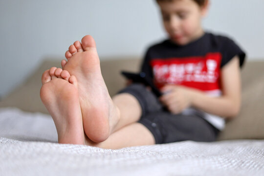 View to bare heels of little boy sitting with smartphone sitting on a sofa. Child playing mobile game or watching cartoons, leisure at home