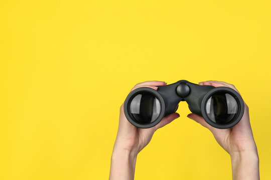Woman holding modern binoculars on yellow background, closeup. Space for text