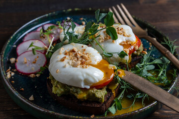 Bread toast, poached eggs, avocado pulp, pea microgreens and fresh vegetables on plate in cafe, breakfast time