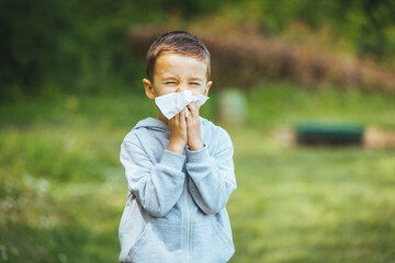 Boy has allergies from flower pollen. Allergy concept. Little boy is blowing her nose near blooming...