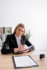 cheerful notary looking at camera while writing in notebook at workplace.