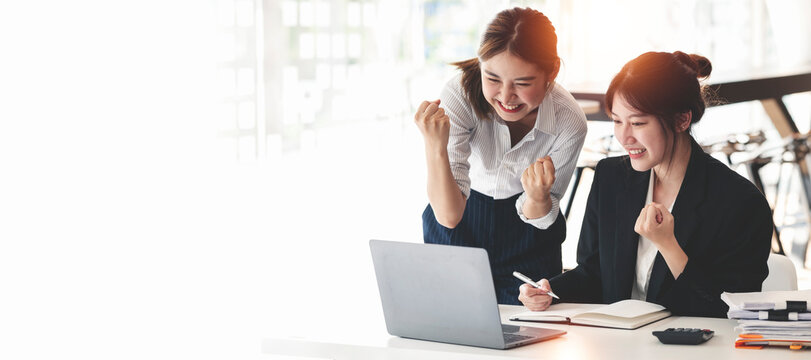 Two Elegant businesswoman sitting in office with laptop. Excited asian business people raising hands to congratulate while working on laptop in office.