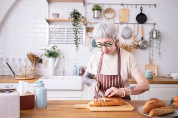 Elder Woman cutting freshly baked bread at wooden table.Retired Asian female Breakfast is being prepared in a cozy kitchen. wearing an apron.person with white hair,gray hair,prepares food for family.