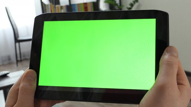 Close-up of hands holding tablet computer with green screen mockup. A man holds a tablet with a green screen on the background of the room. Template for video conference.