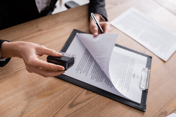 cropped view of lawyer holding stamper and pen near contract on clipboard.