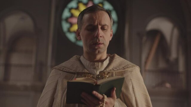 Chest up slowmo of Caucasian priest in white vestment reading prayer and crossing standing in dark Catholic church with beautiful round stained glass window behind