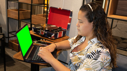 joven mujer sentada frente a computadora junto a tornamesa con disco de vinil, pantalla verde en la lap top