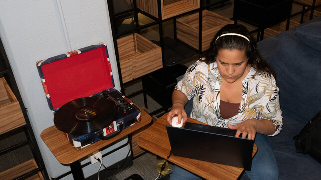 senital joven mujer sentada en sill&oacute;n junto a tornamesa con disco de vinil trabaja en casa con laptop mira a la computadora 