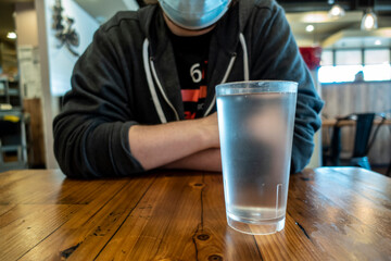Low angle view of a man in a blue face mask, sitting at a wooden table with a glass of water inside a restaurant