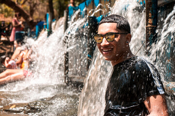 Fototapeta premium Latino teenager in sunglasses smiling bathing in a river with the water from a dam falling on his back