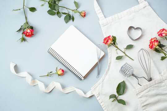 Blank recipe cooking book, baking accessories with apron with flowers roses on a gray background, flat lay, top view. Festive baking recipes concept