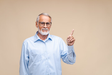 Fototapeta premium Elderly businessman with happy face pointing up with finger at copy space. Indoor studio shot isolated on beige background