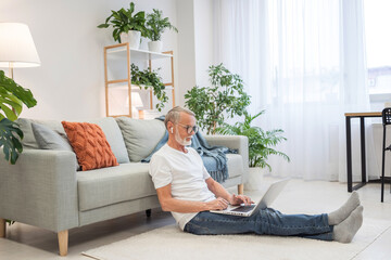 Mature grey-haired male entrepreneur using laptop at the floor at home, takes notes, multitasking senior good looking man working remotely from home