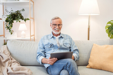 Cheerful senior man wearing casual wear sitting on the sofa and using laptop at modern home office, glad male entrepreneur typing on keyboard