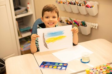 Cute boy smiling while sitting at his desk at home and holding a drawn flag of Ukraine.
