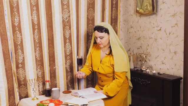 A Jewish woman in a wig and head covering makes a celebratory kdush with a glass in her hand and a siddur. Overall plan