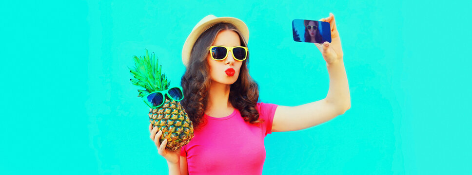Summer portrait of young woman with pineapple taking selfie by smartphone and blowing her lips wearing straw hat, sunglasses on blue background