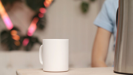 Close-up of white cup and electric kettle standing on the wooden table and silhouette of young...