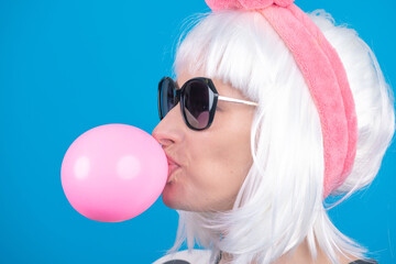 Fashion, style and makeup concept. Close-up studio portrait of beautiful woman with big sunglasses and white bob hairstyle wig bound with pink fluffy headband blowing pink chewing gum bubble