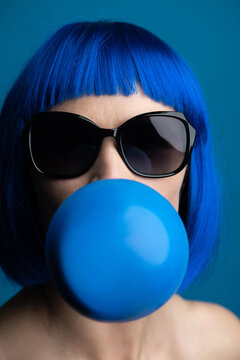 Fashion, style and make-up concept. Close-up studio portrait of beautiful woman with big sunglasses and blue bob hairstyle wig blowing blue chewing gum bubble with copy-space. Blue studio background