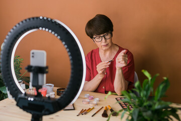 Middle-aged woman talking on cosmetics with makeup eye shadows and blush palette and brushes while recording her video. Mature female making video for her blog on cosmetics