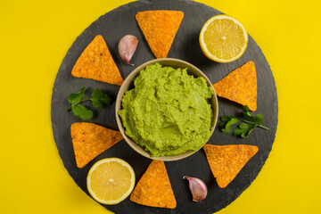 Composition of guacamole with the ingredients for its creation and the nachos for its consumption on a yellow background.