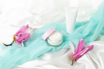 Fototapeta na wymiar cream jar and plastic container mockup with magnolia flowers on white satin background