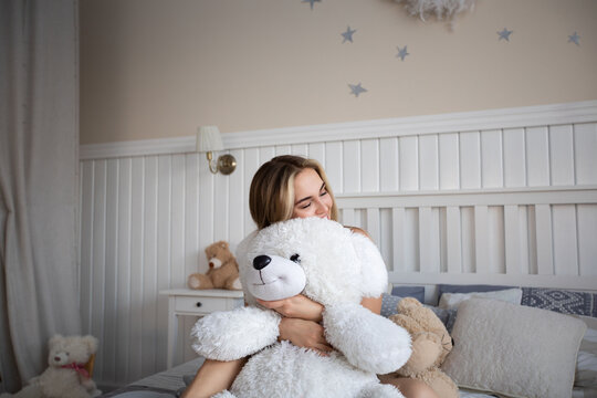 Cute blonde woman sitting with teddy bear on bed in bedroom.