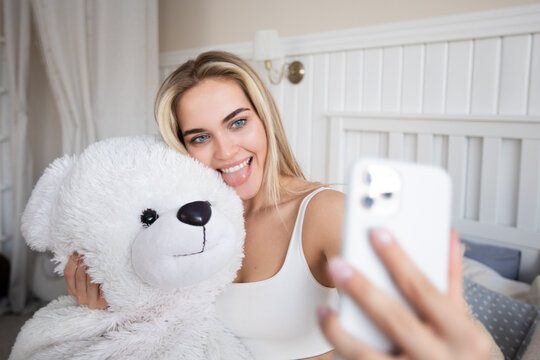 Beautiful blonde woman makes a selfie with a teddy bear sitting on the bed.