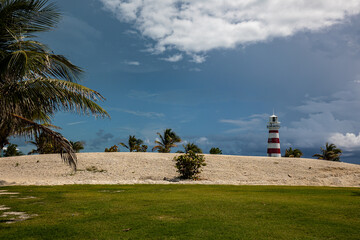 A bank of sand behind grass lawn and in front of a lighthouse on a tropical island
