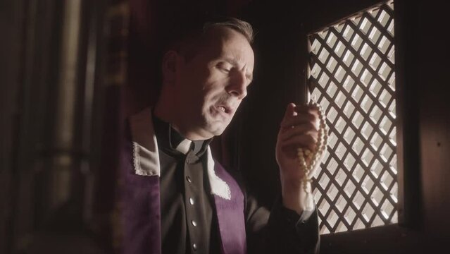 Waist up slowmo of mature priest wearing black and purple robe talking to parishioner while sitting in dark confession booth with rosary beads in hands