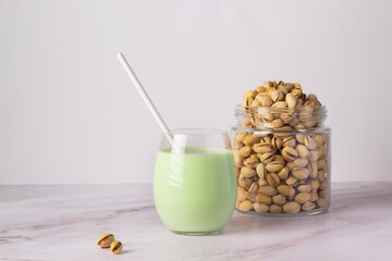 Clear glass filled with creamy pistachio milk, accompanied by a straw and a full jar of shelled pistachios on a clean marble surface.
