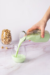 Pouring vibrant pistachio milk from a glass bottle into a clear tumbler, with a nearby container of shelled pistachios on a pristine marble backdrop.