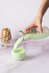 Hand pouring creamy pistachio milk into a transparent glass, set against a white marble backdrop with whole pistachios in a jar nearby.