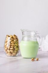 Jug filled with fresh pistachio milk stands next to a jar of whole pistachios, with a few nuts scattered on a marble counter.