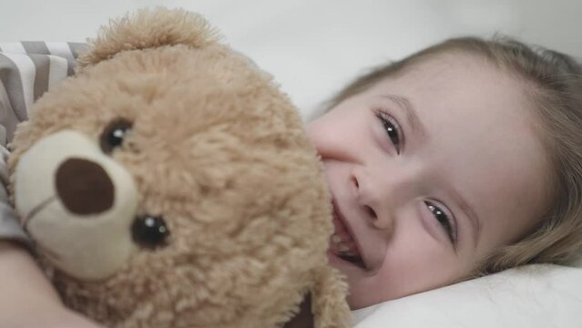 Small child girl smiles lying on bed and hugging teddy bear