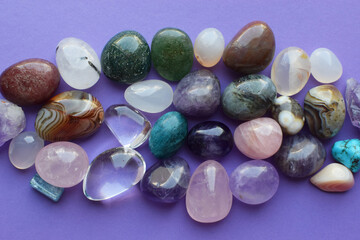 Tumbled gems of various colors. Amethyst, rose quartz, agate, apatite, aventurine, olivine, turquoise, aquamarine, rock crystal on purple background.