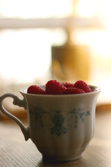 Porcelain cup filled with raspberries, open book, reading glasses and vase with flowers on the table. Selective focus.