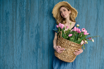Fototapeta na wymiar beautiful young florist with a wicker basket of flowers in her hands and a straw hat on a blue background