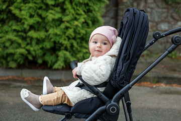 Little very beautiful girl is sitting in stylish black stroller. Gently pink hat and beige jacket