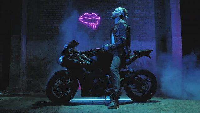 A man with a bandama sits on a sports motorcycle and texts on a mobile phone against a brick wall with a neon sign and smoke