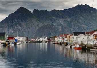 Fototapeta na wymiar Norwegian seascape, cityscape of the town Henningsvaer, a small boat moves between peninsulas, sail boat, rocky coast with dramatic skies, classic view of Norwegian houses on the slopes