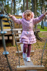 Cute happy little girl in winter faux fur ear muffs plays outdoors at playground on warm sunny autumn day - 502481864